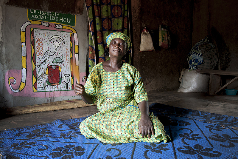 Marguerite Adjahi s'occupe de ses frères et beaux frères quand ils dansent en coulisse, jouant ainsi de manière informelle le rôle de 'Iyaidjo'. Elle affirme que ses ancêtres font partie de ceux qui ont amené la danse Guèlèdè d'Ilobi au Nigéria jusqu'à Ofia. Ofia, Bénin, 2017. ©Laeïla Adjovi